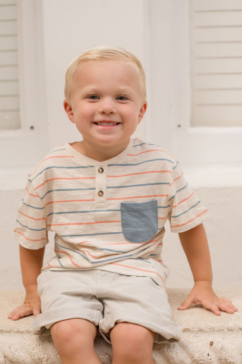 A young child in a striped shirt and shorts sits on outdoor concrete steps with white shutters in the background.