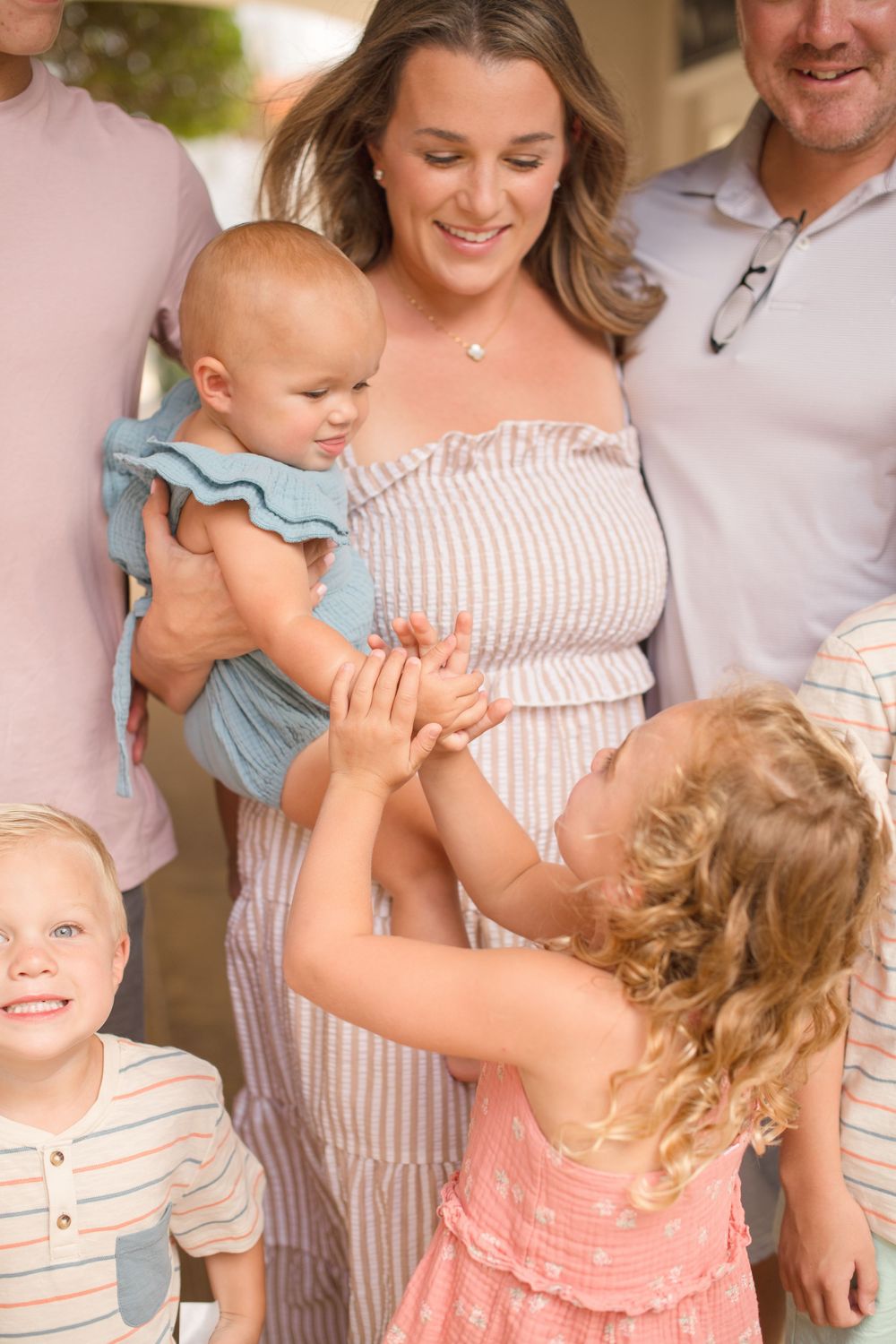 Sweet family moment shows baby in blue outfit being lifted up while wearing a striped dress and pink sundress.