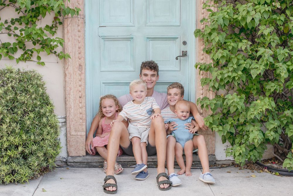 Children sit together in front of a blue door with greenery around it creating a charming summer scene.
