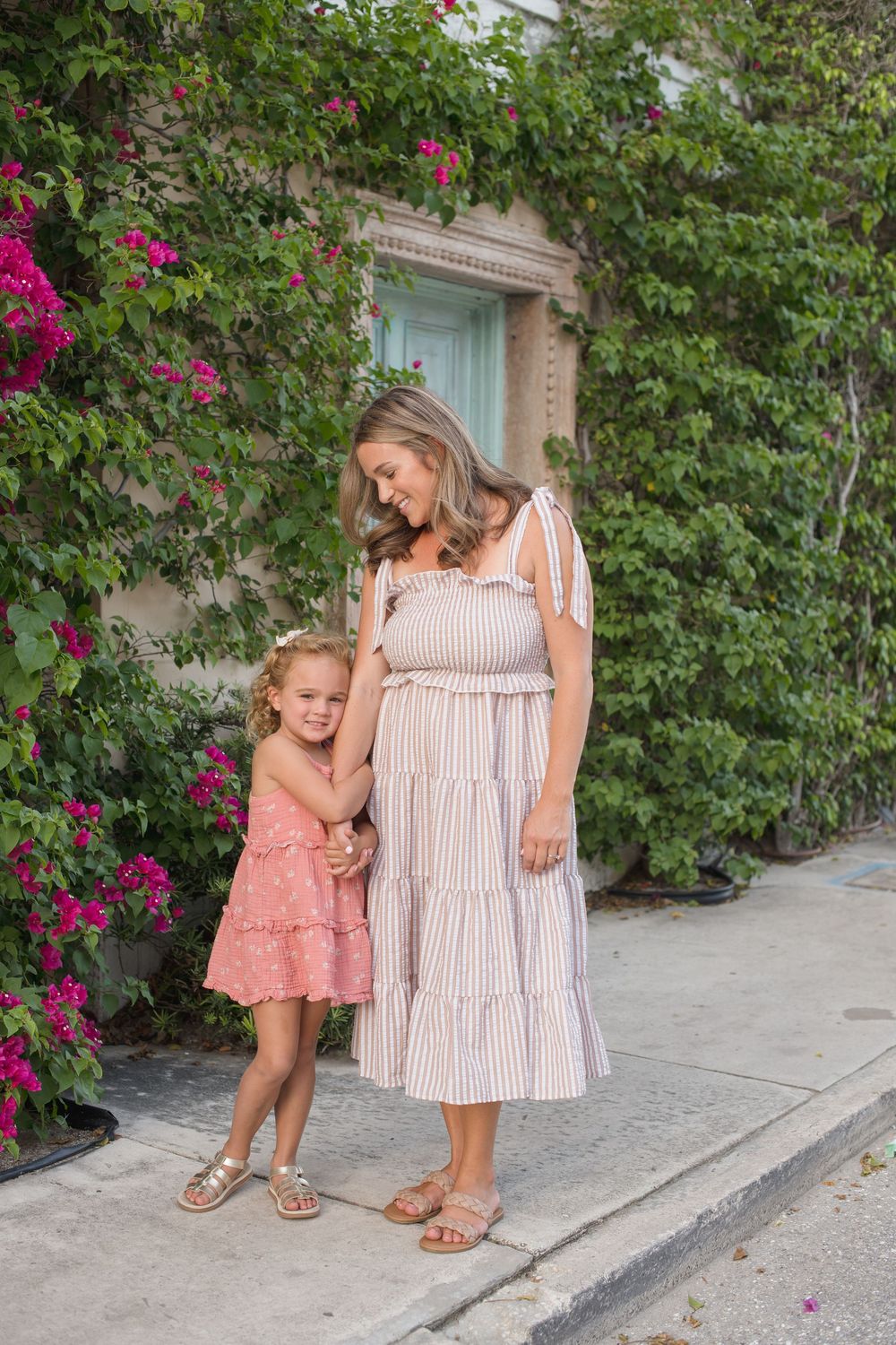 Pair wearing coordinating summer dresses stand together beside blooming pink bougainvillea and garden path.