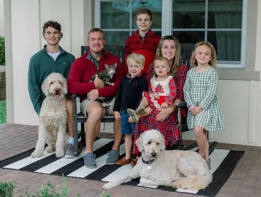 Group sitting together with two goldendoodle dogs on a black and white striped outdoor rug by a front porch.