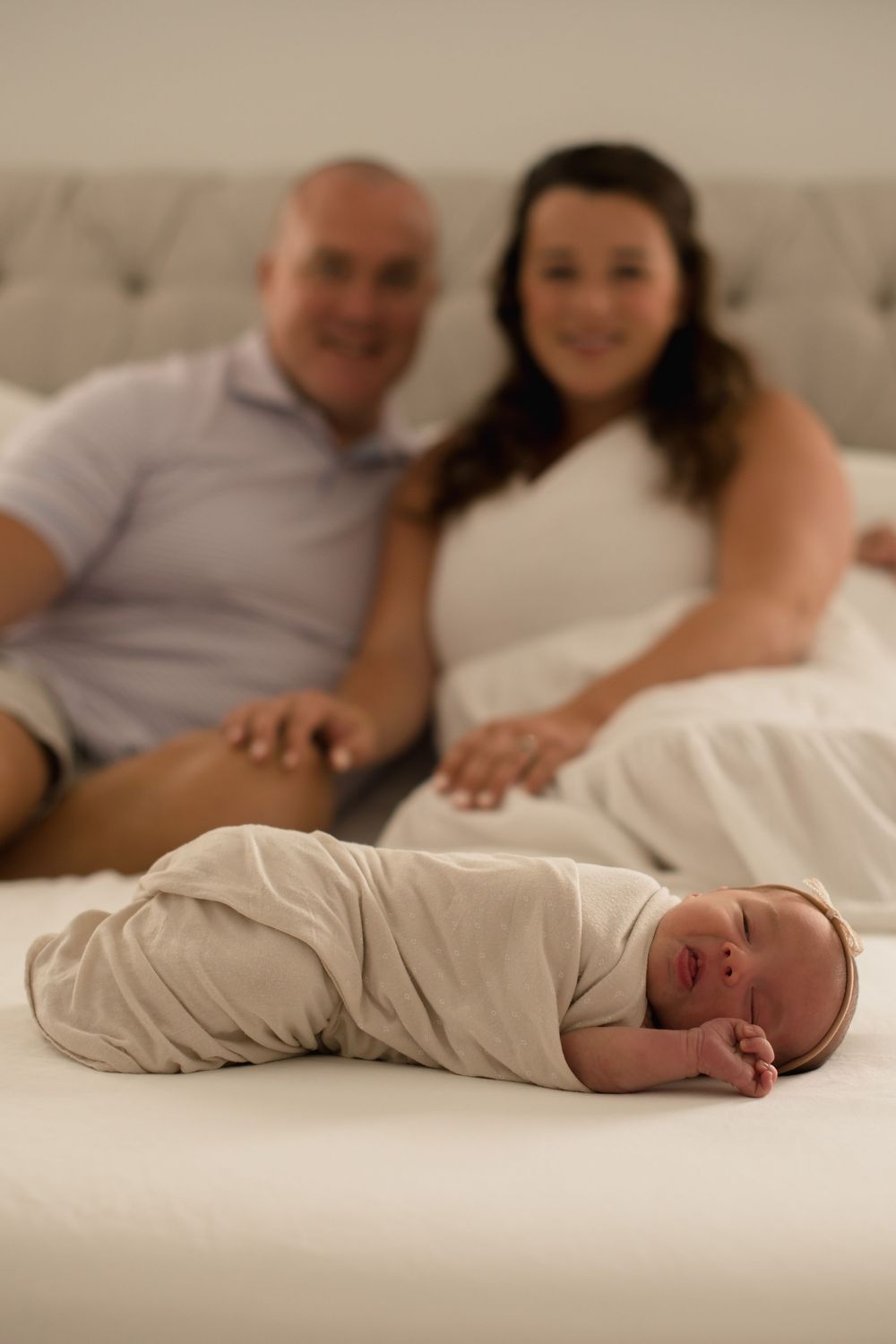 A swaddled newborn baby lying on a white couch while parents sit in the background.