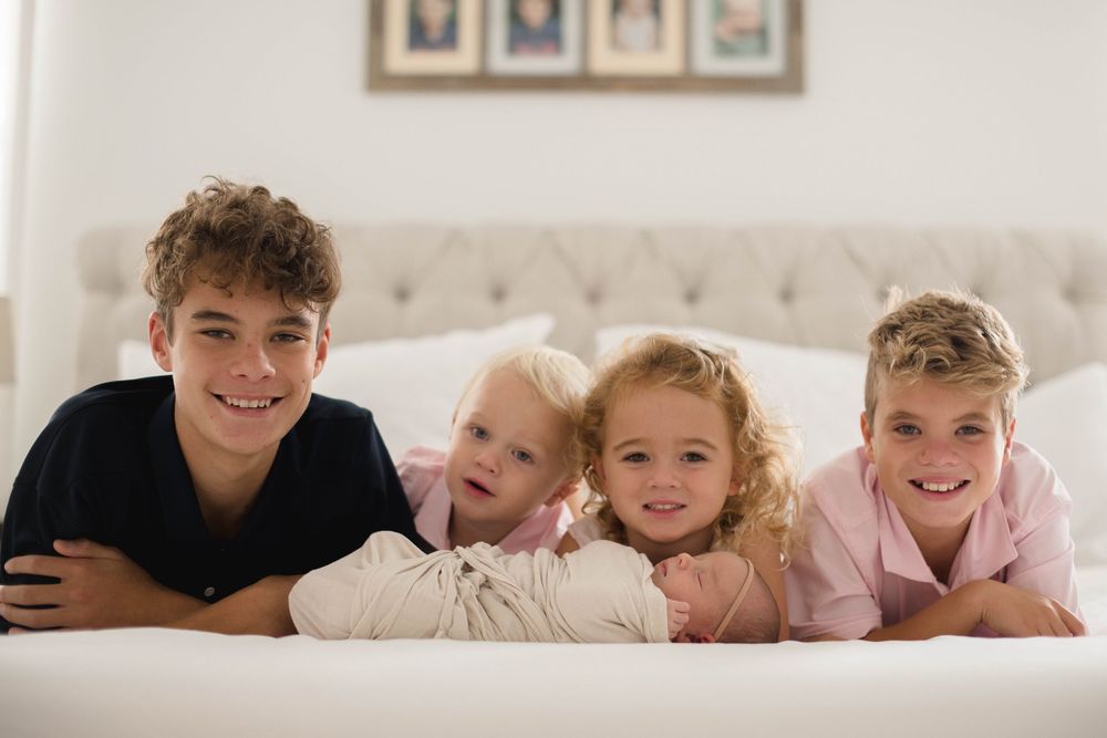 Four siblings laying together on a white bed wearing pink and black casual clothing.