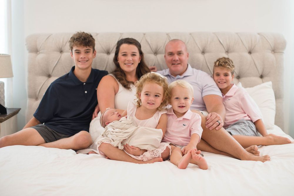 A family lounges together on a white bed wearing casual clothing while smiling at the camera.