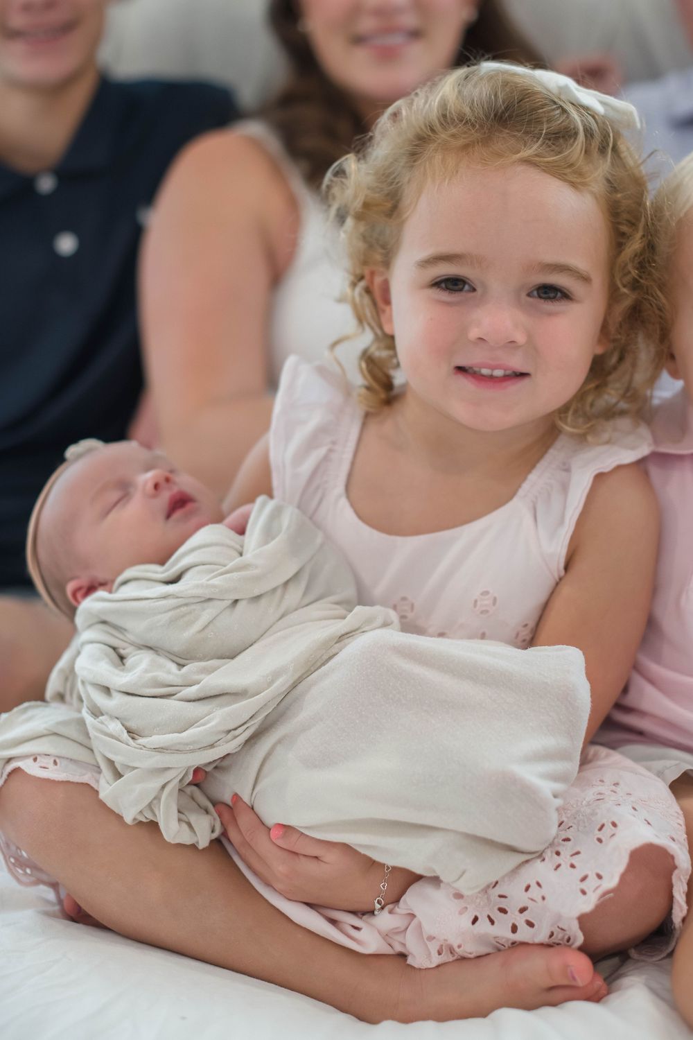 A young curly-haired sibling in a white dress holds a swaddled newborn baby while sitting together.