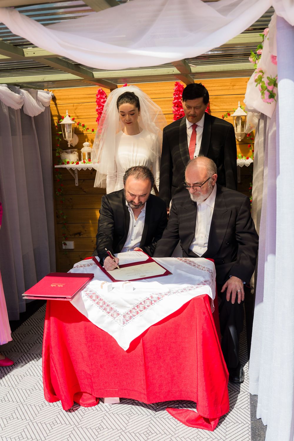 Wedding ceremony participants signing documentation at a table with red tablecloth under white draped canopy.