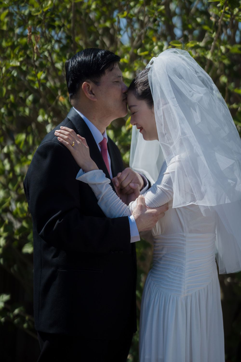 A newlywed couple shares an intimate moment outdoors with the bride in a white gown and veil and groom in dark suit.