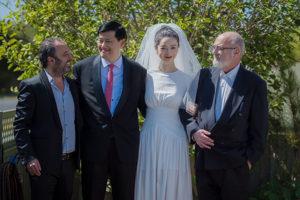 Group in formal wedding attire posing together outdoors in front of greenery and sunlit garden.