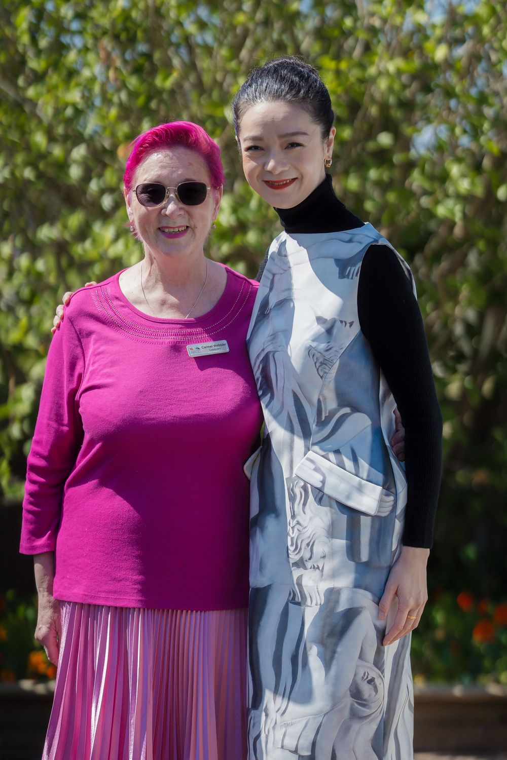 Two people in contrasting outfits pose together outdoors against a green leafy background.