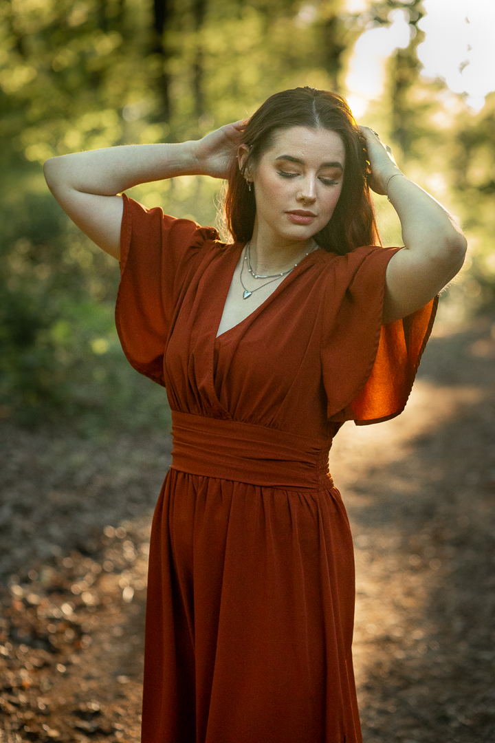 Vrouw in een roest kleur jurk poseert in het bos, handen in het haar.