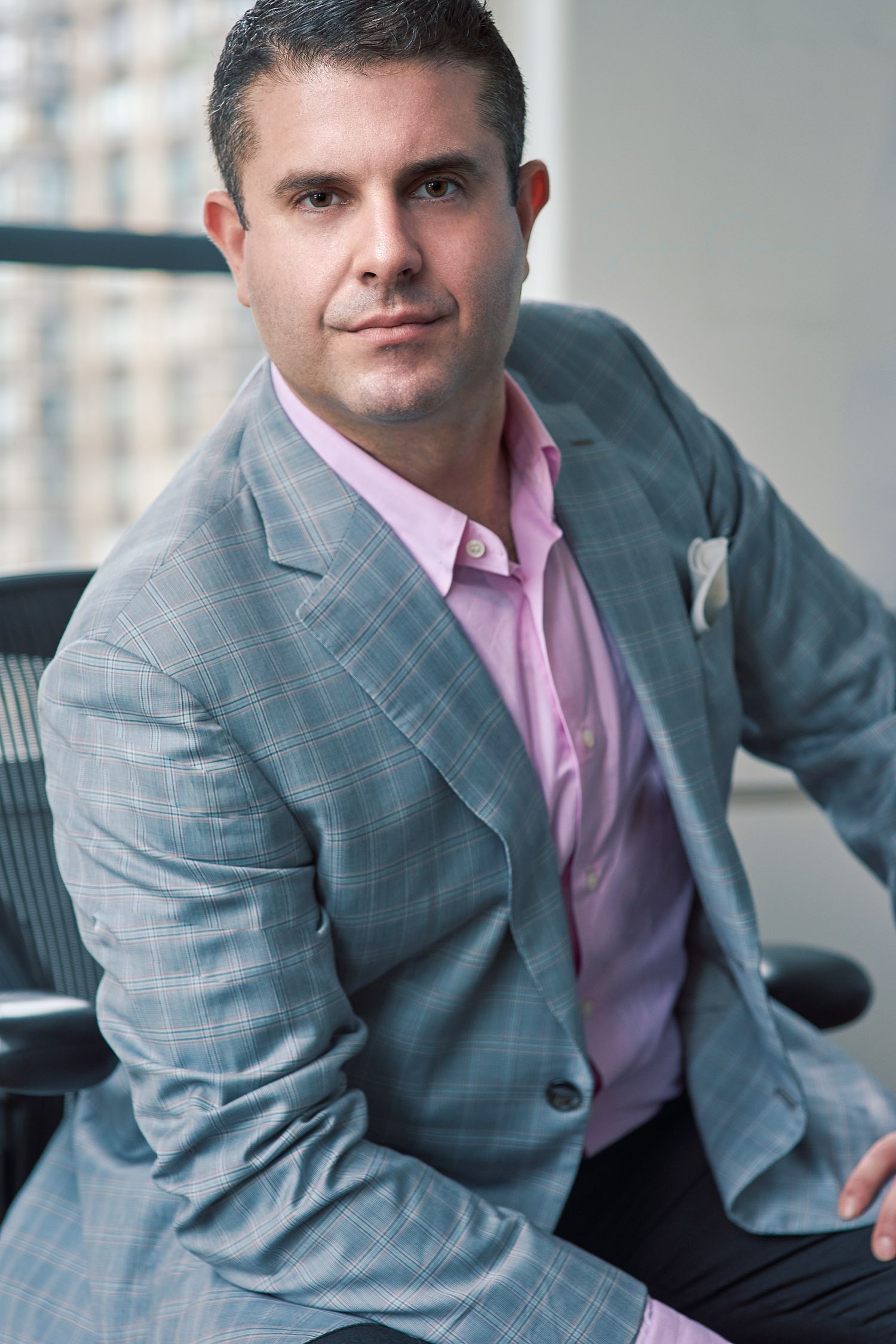 Professional man in gray plaid blazer and pink shirt seated confidently in modern office setting.
