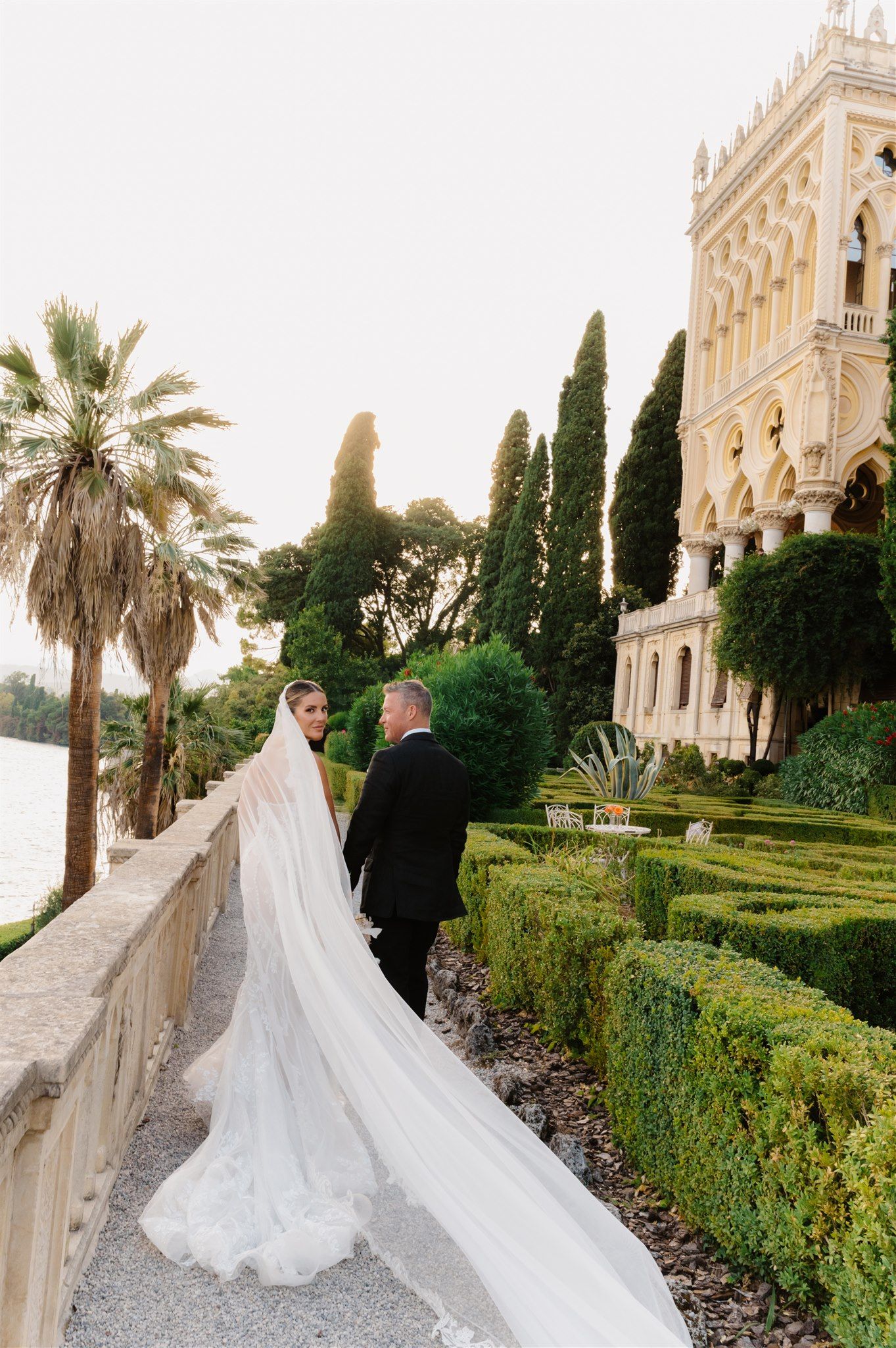 Couple walks through manicured garden path with bride's long veil trailing behind them.