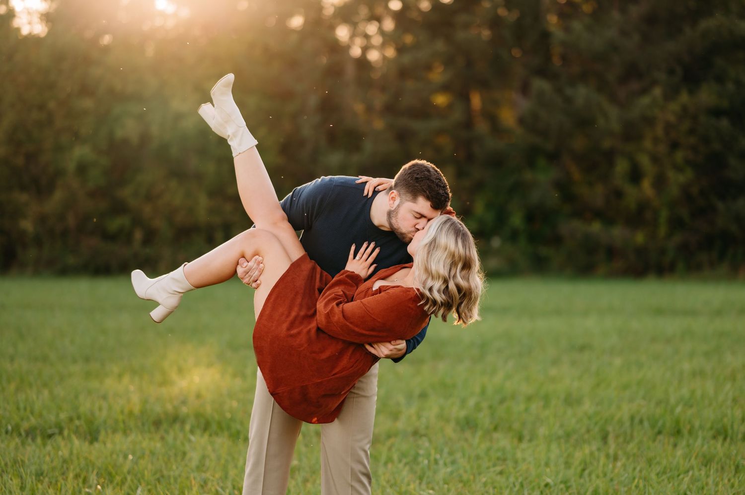 Playful couple in casual clothes doing romantic dip pose in sunlit meadow at golden hour.