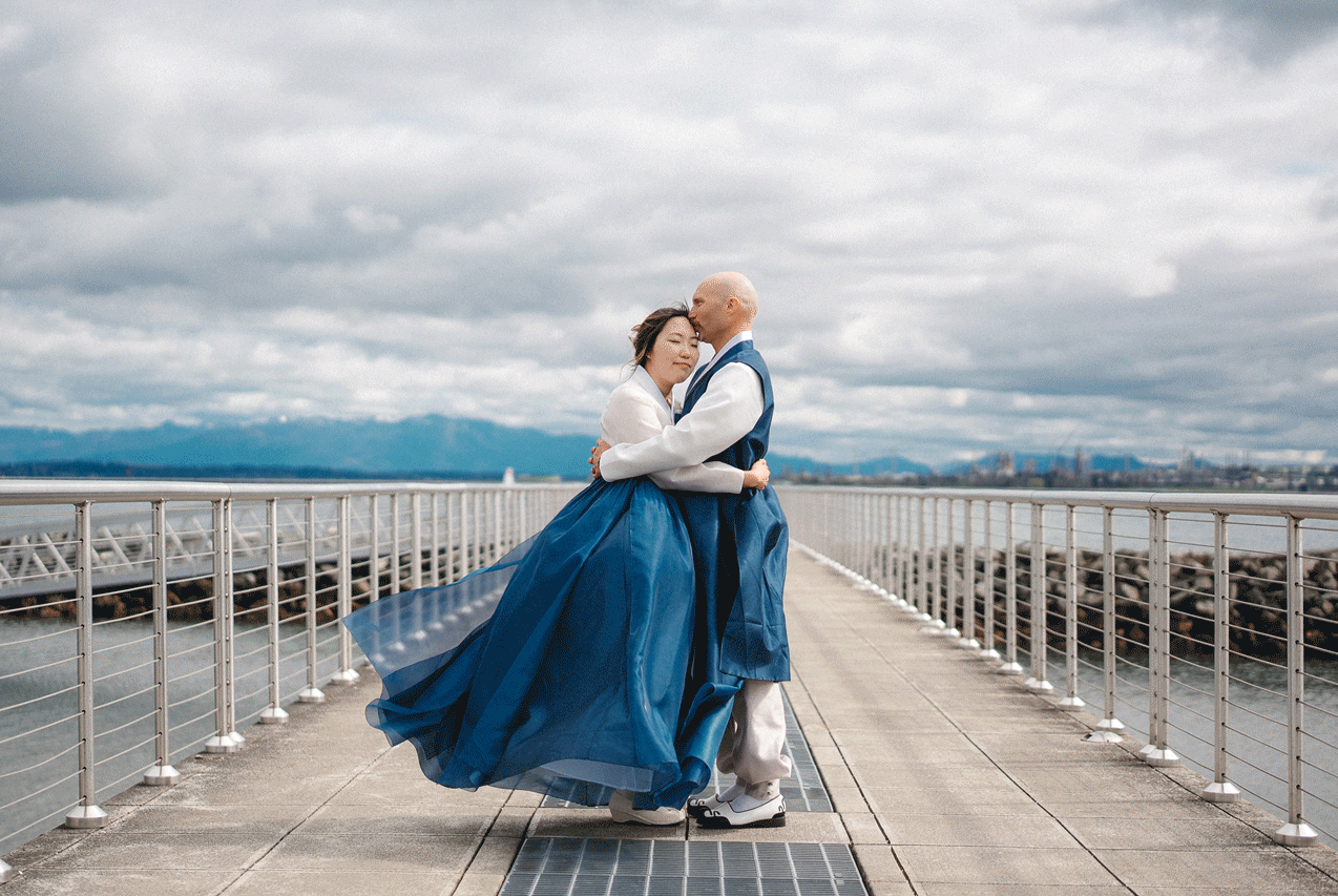 Couple in elegant attire embrace and dance on a waterfront pier under dramatic cloudy skies.
