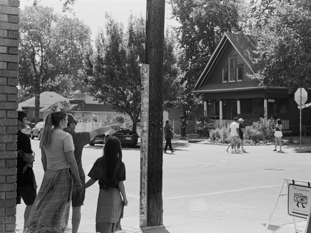 Vintage black and white street scene showing people waiting at a corner with houses and trees visible in the background.