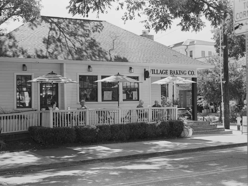 Black and white photograph of Village Baking Co storefront with white picket fence and trees along a street.