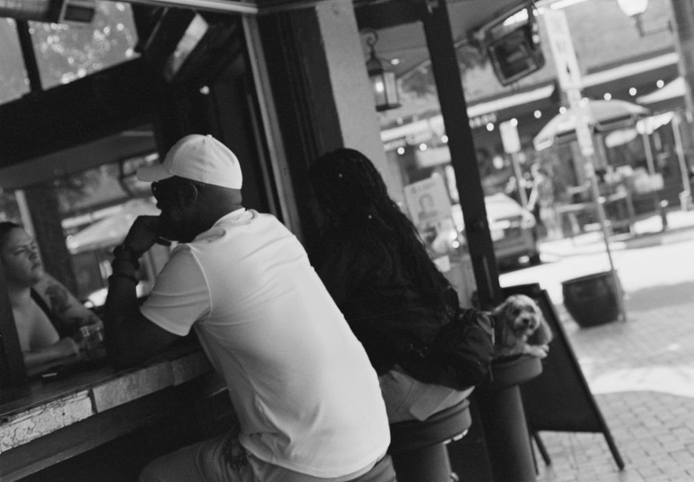 A black and white photograph showing a person in a white t-shirt and baseball cap leaning against a counter at a street corner.