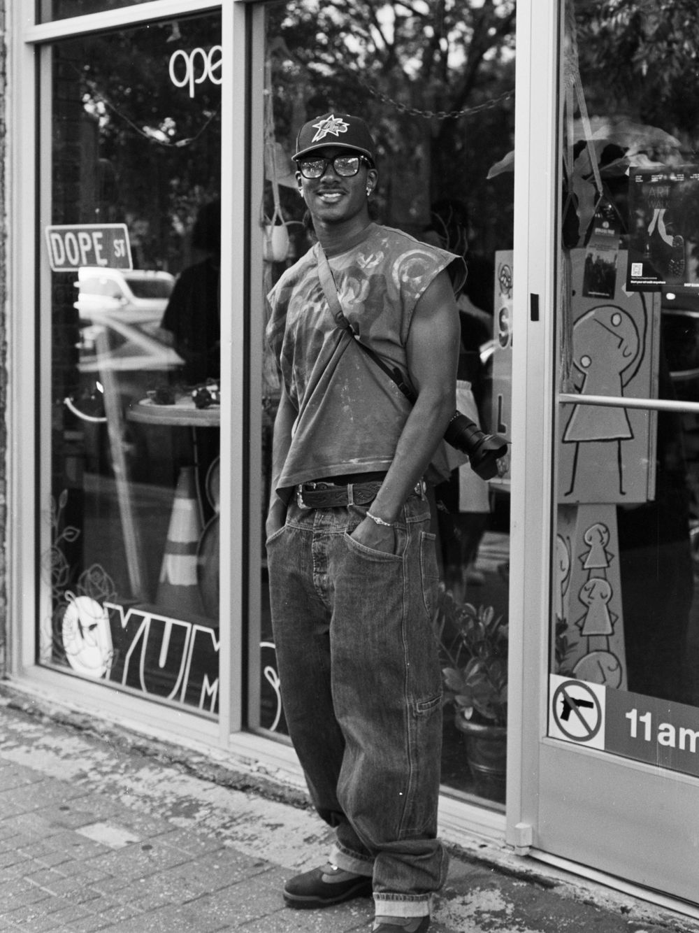 A black and white photo of someone standing in front of a storefront window wearing casual clothes and a baseball cap.