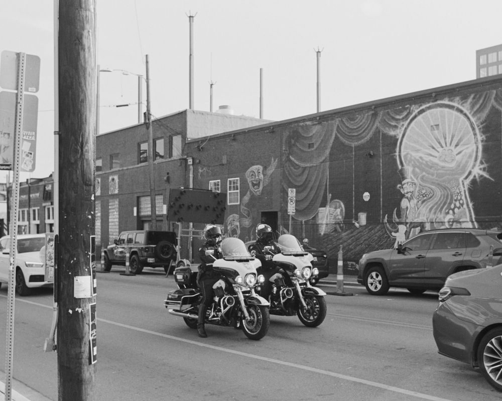 Two police motorcycles parked on a city street with a colorful mural on the brick wall behind them.