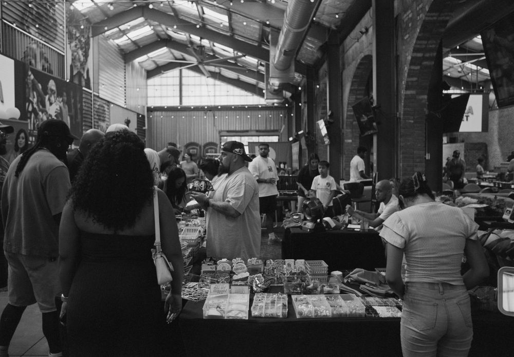 Group of shoppers browse through items at an indoor market with dark lighting and crowded walkways.
