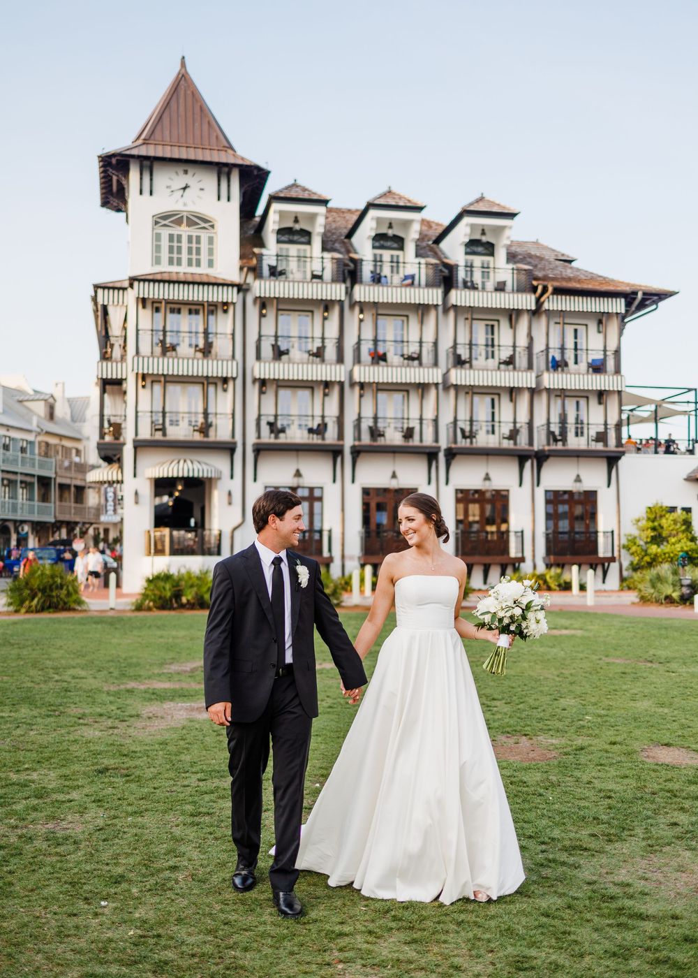 A bride and groom walk together on the lawn of a elegant Victorian-style venue with white balconies and brown roofs.