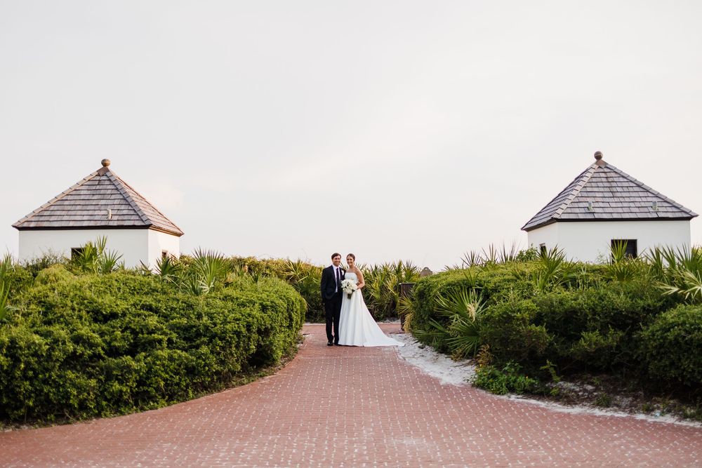 A couple stands between two white gazebos on a brick path surrounded by manicured hedges on a cloudy day.