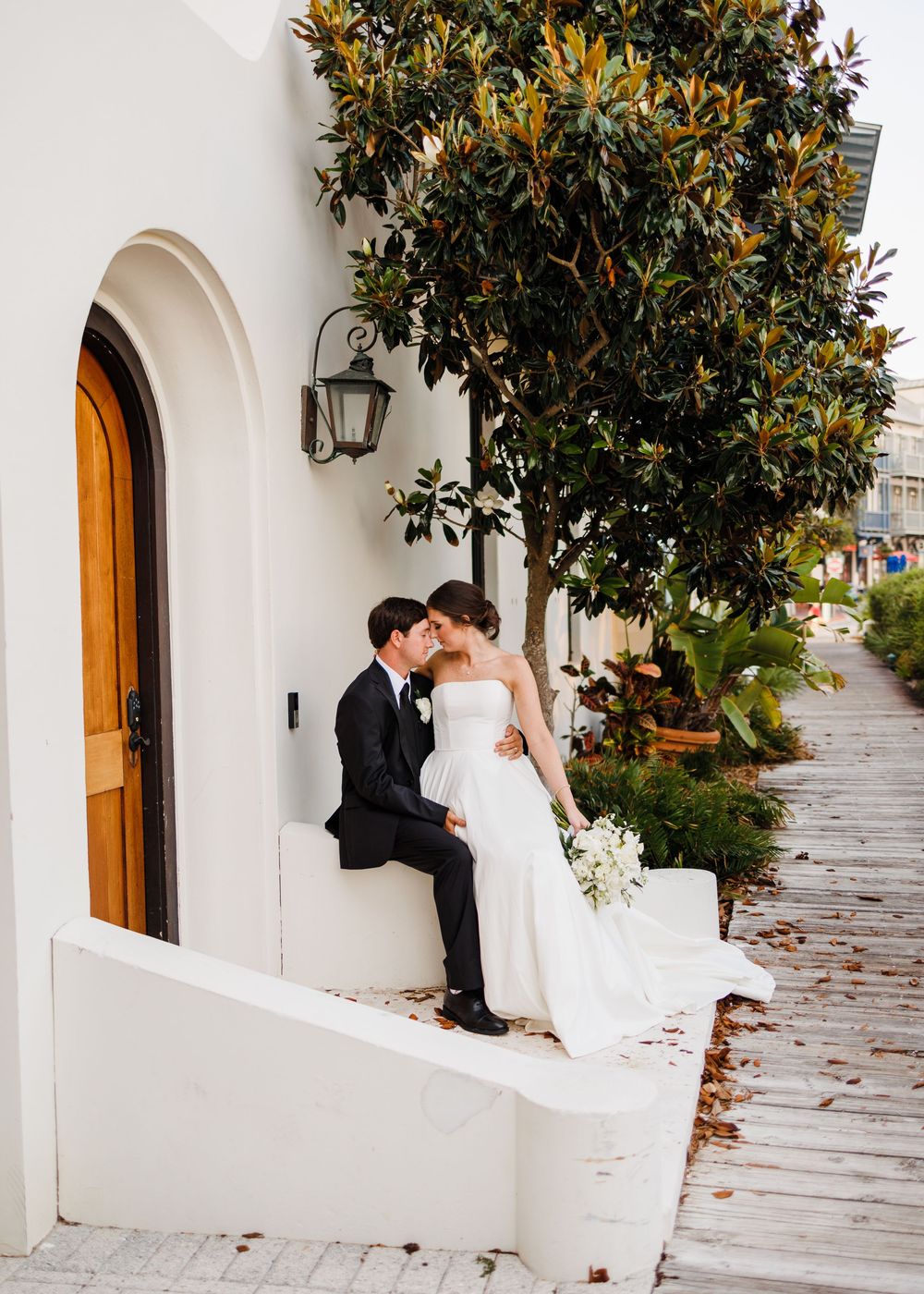 A romantic couple in formal wedding attire share an intimate moment on a white-walled patio with arched doorways and magnolia trees.
