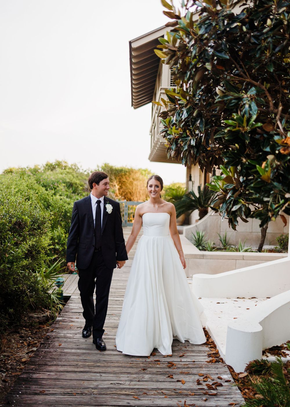 Newlywed couple walking together on a wooden pathway surrounded by lush greenery and magnolia trees on a sunny day.