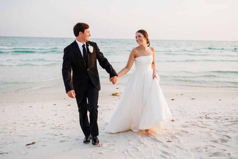 Couple in wedding attire walks together along a serene white sand beach at sunset with ocean waves in the background.