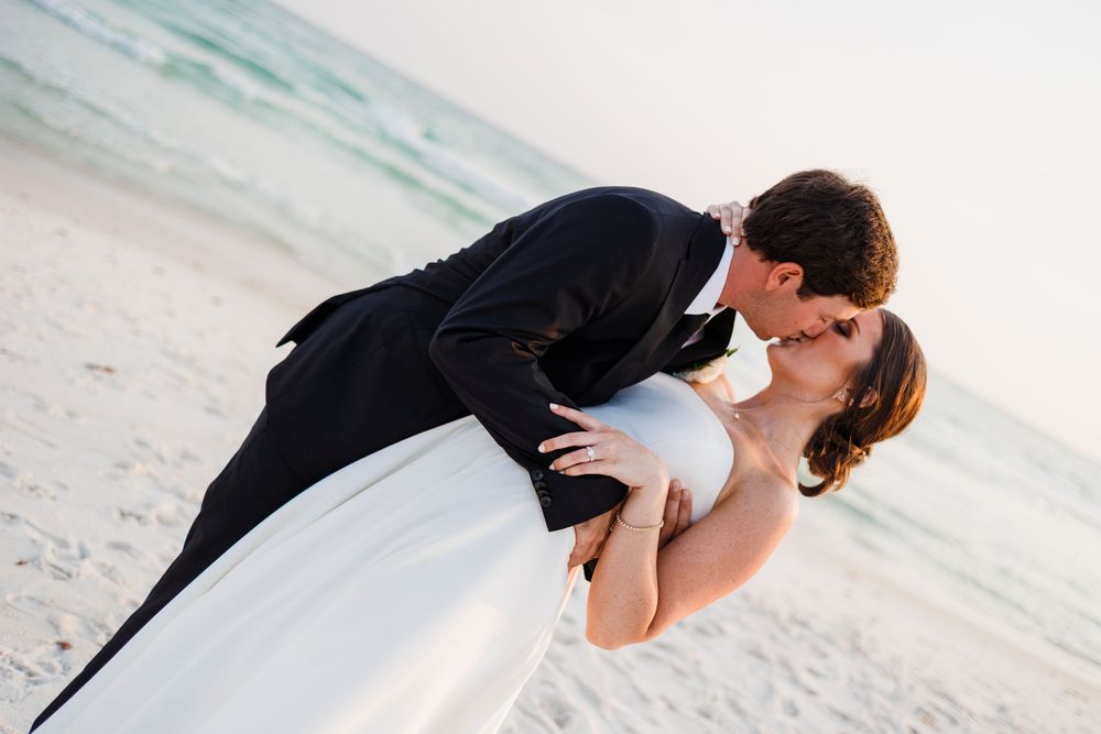 A couple in wedding attire share a romantic kiss while embracing on a beach at sunset.