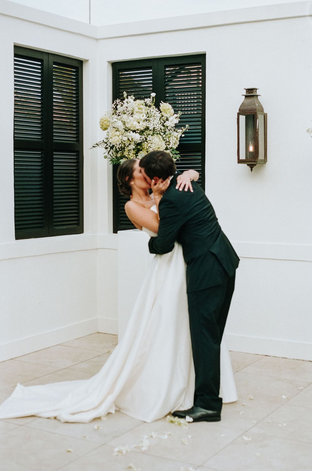 A romantic couple embracing on their wedding day against a white wall with green shutters and a vintage lantern.