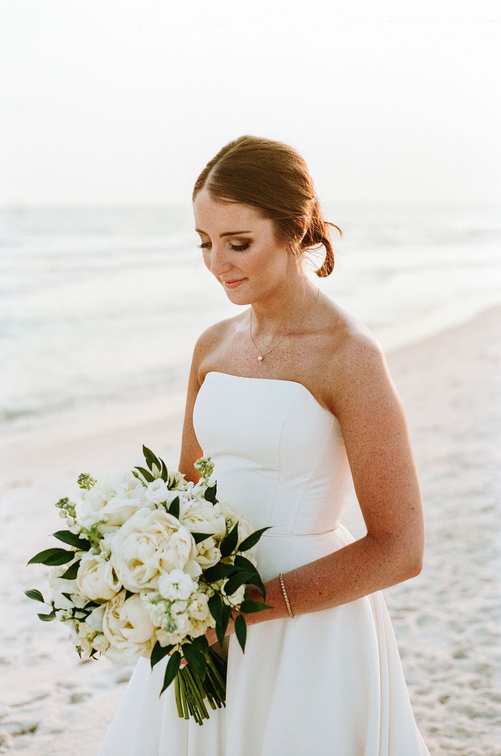 Beautiful bride in a strapless white dress holds elegant white floral bouquet on a serene rosemary beach at sunset.