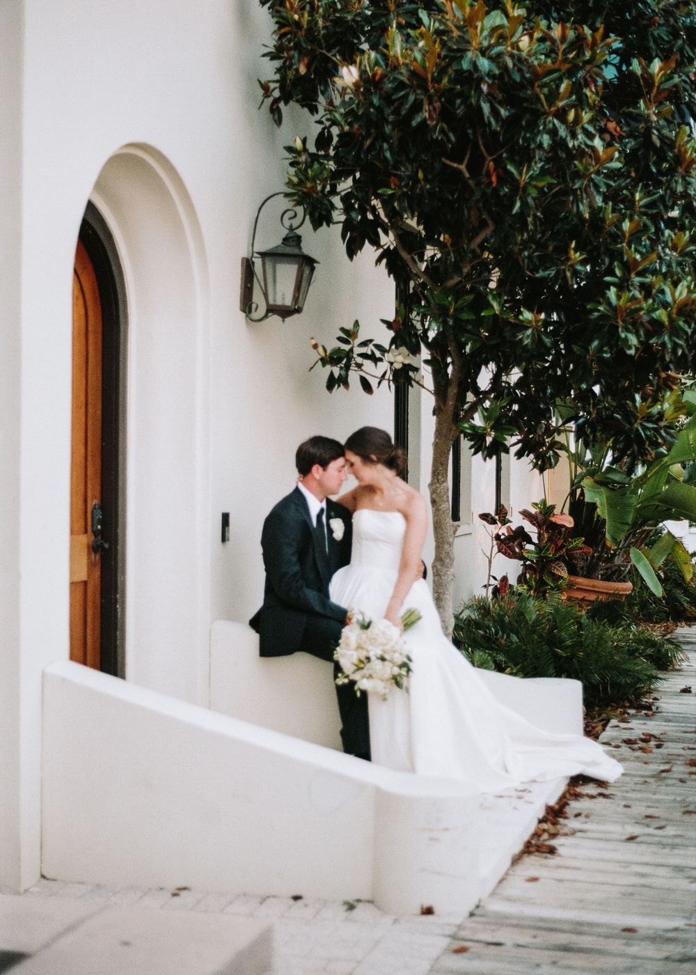 A tender wedding moment captured on a white-walled terrace with an arched doorway and lush greenery overhead.