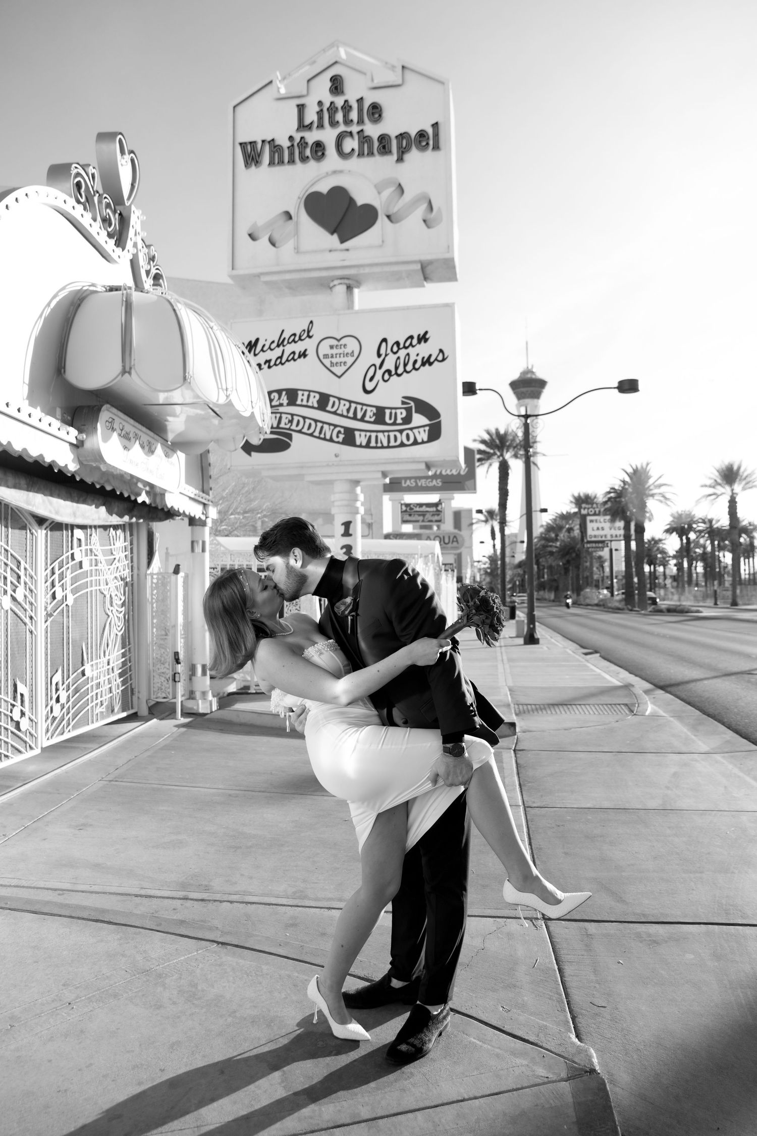 A romantic couple embraces outside the Little White Chapel wedding venue in Las Vegas, with palm trees visible in the background.