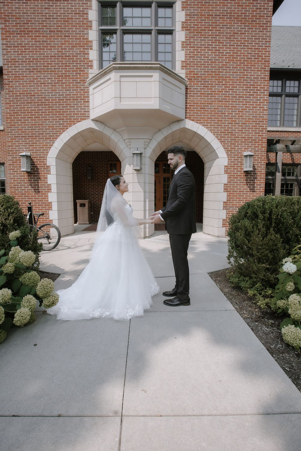 Bride and groom stand beneath an elegant brick archway entrance with decorative hydrangea bushes on a sunny day.