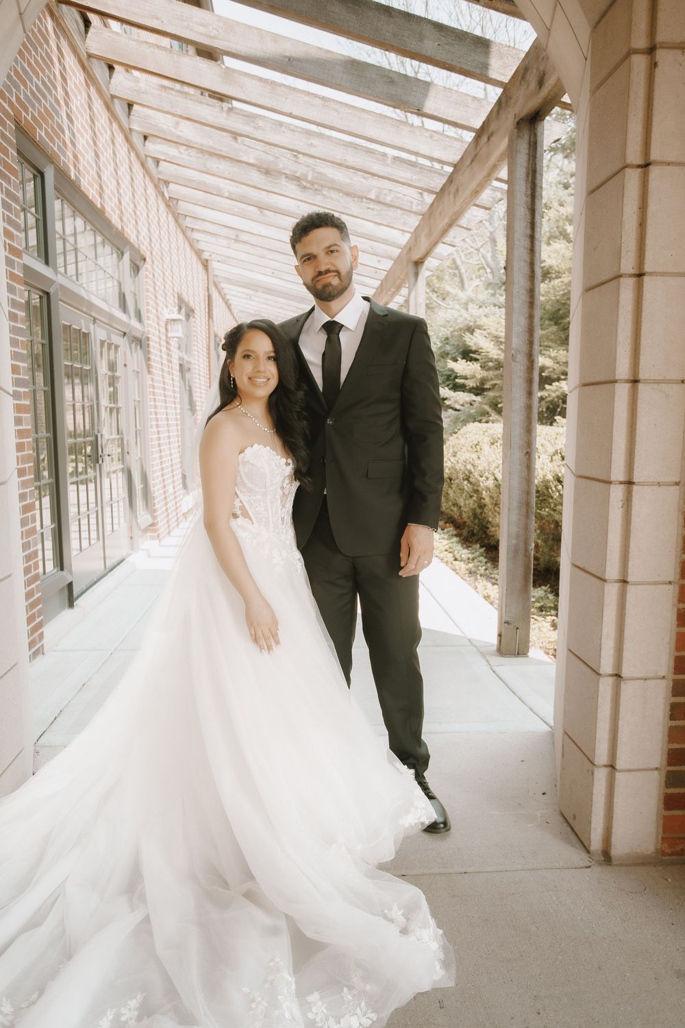A bride in a flowing white wedding gown and groom in black suit pose under a covered walkway on their wedding day.