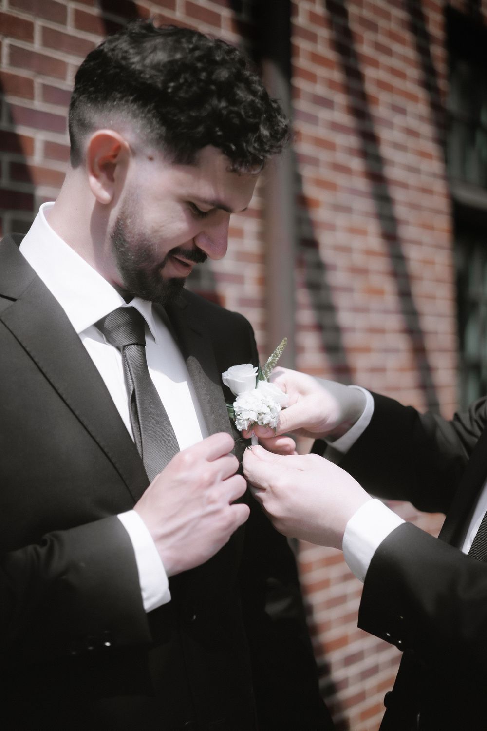 Groom in dark suit getting boutonniere pinned during wedding preparations against brick wall.