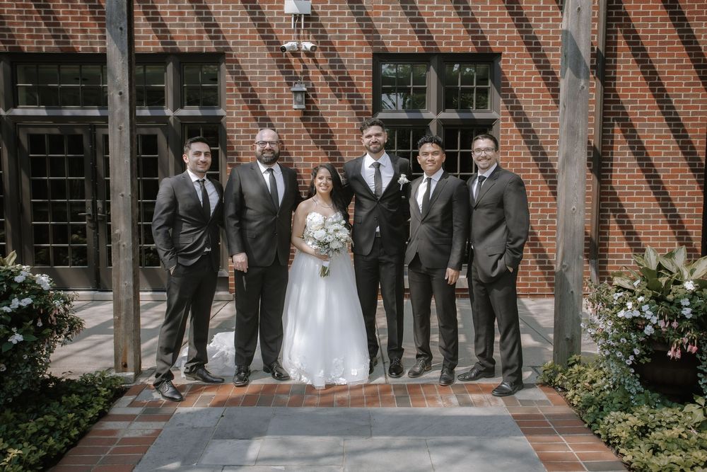 Wedding party with groomsmen in grey suits lined up against red brick exterior.