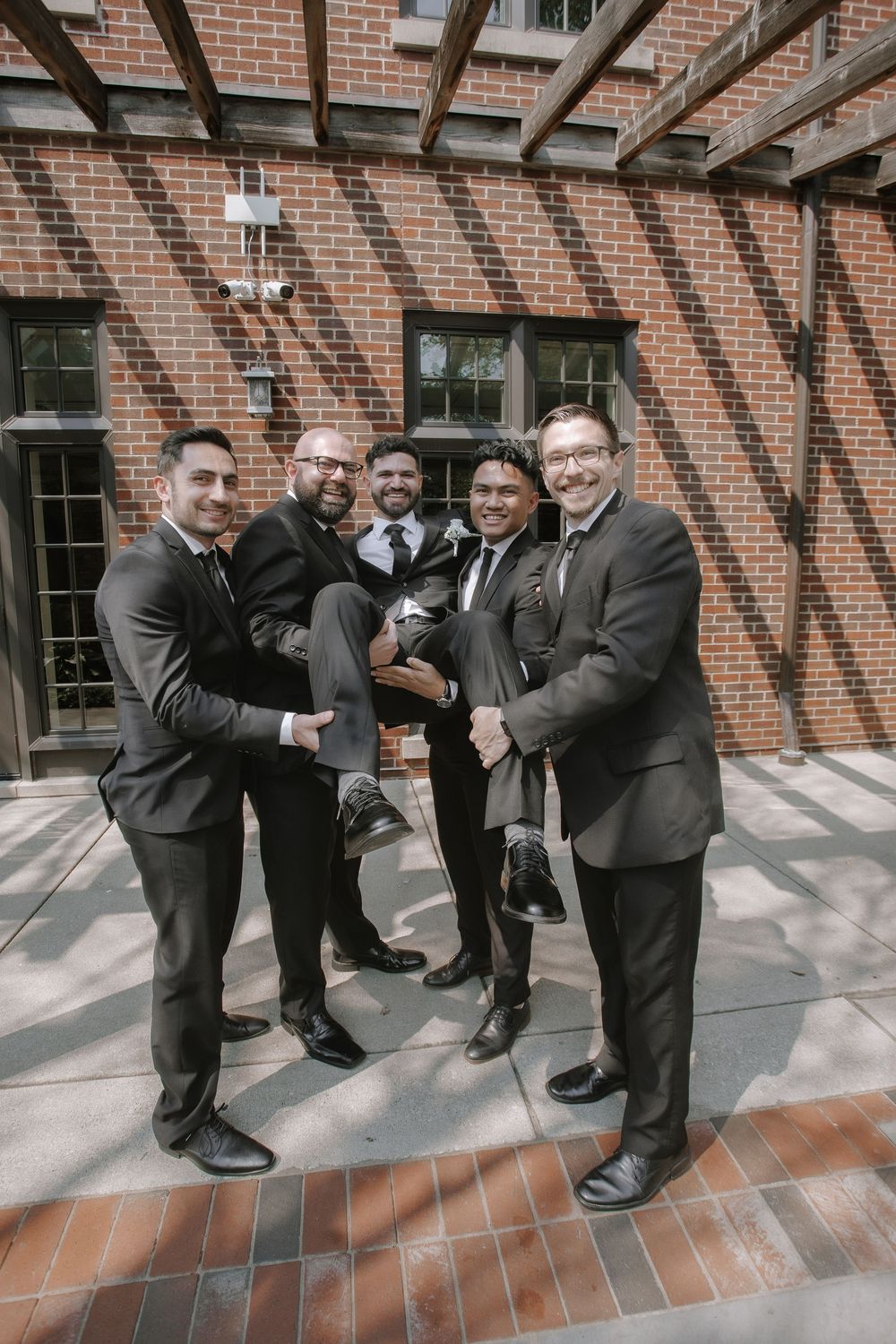 A sequence of groomsmen posing playfully in black suits near a brick wall with shadowed arches.
