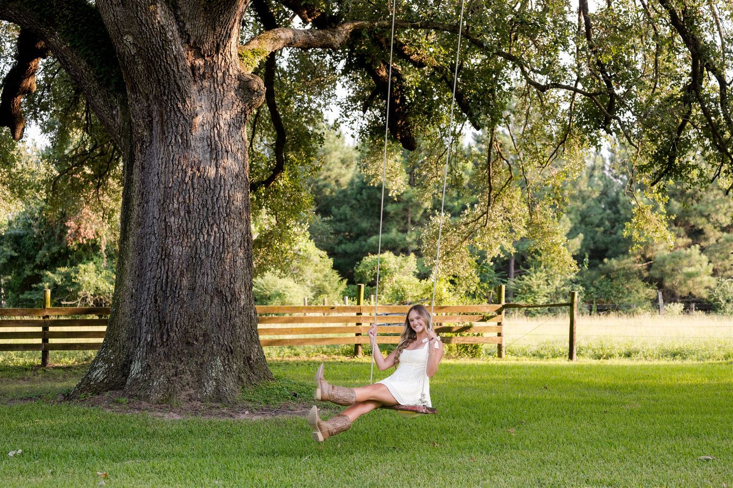 A high school senior girl in a flowing yellow dress dances gracefully near a large oak tree in a sunlit farm setting.