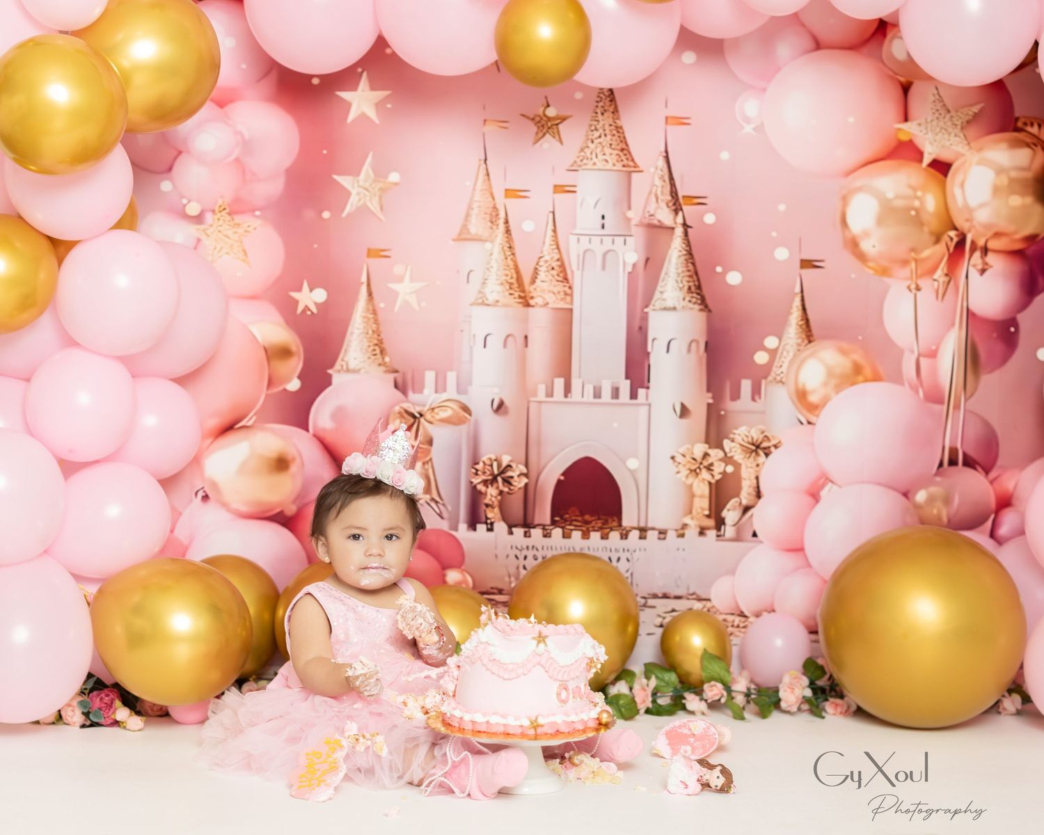 Elegant pink and gold balloon garland backdrop with castle decorations and birthday cake for a baby's first birthday celebration.