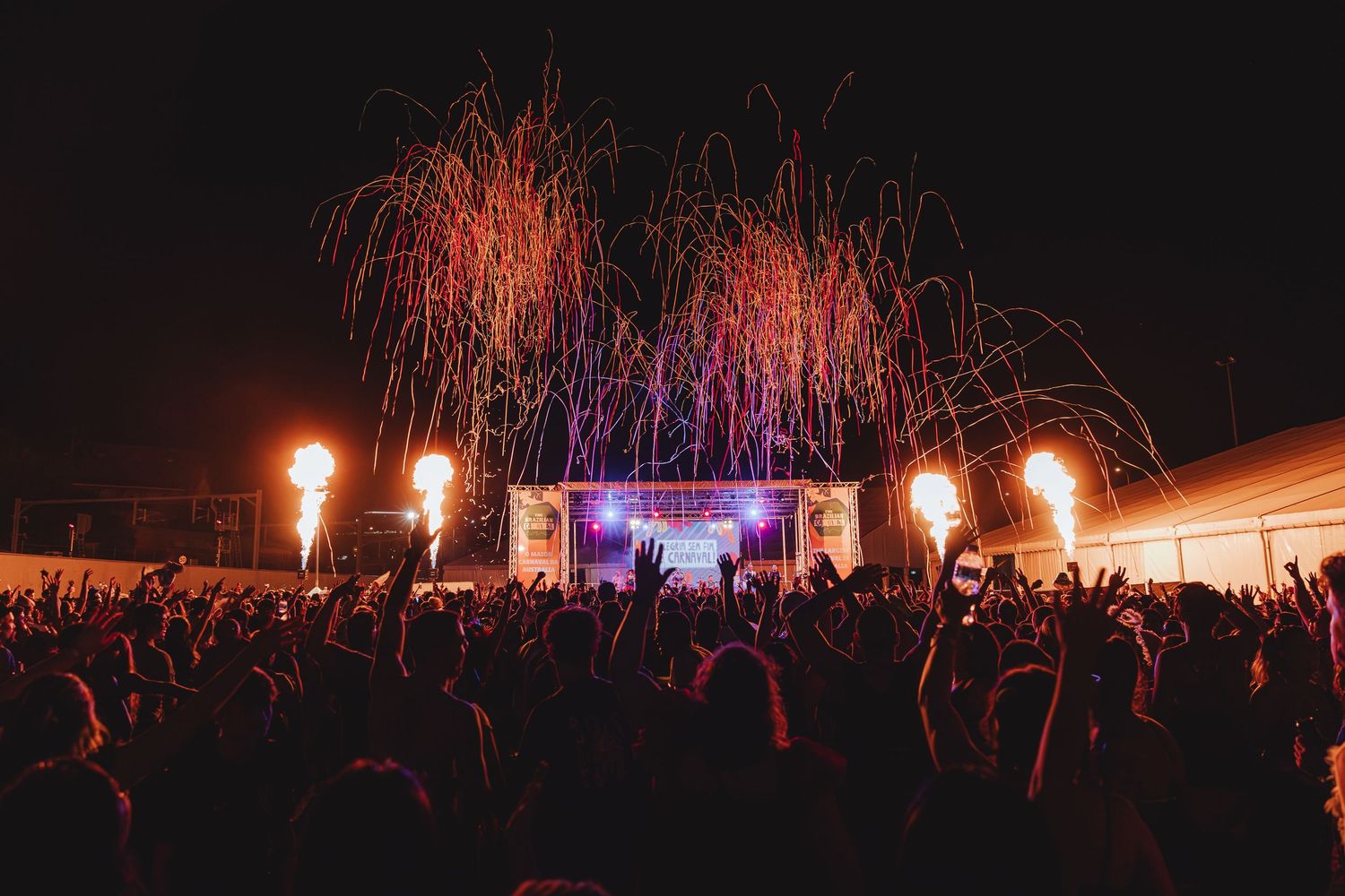 Large crowd at indoor concert venue with bright pyrotechnic flames illuminating the stage and audience in orange glow.