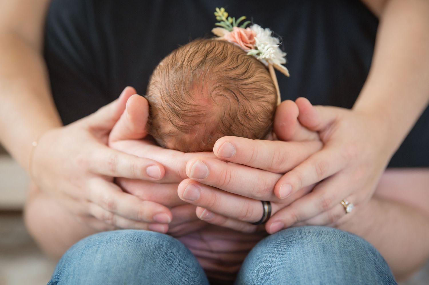 Gentle hands cradle a newborn's head adorned with a delicate floral headband while sitting in denim jeans.