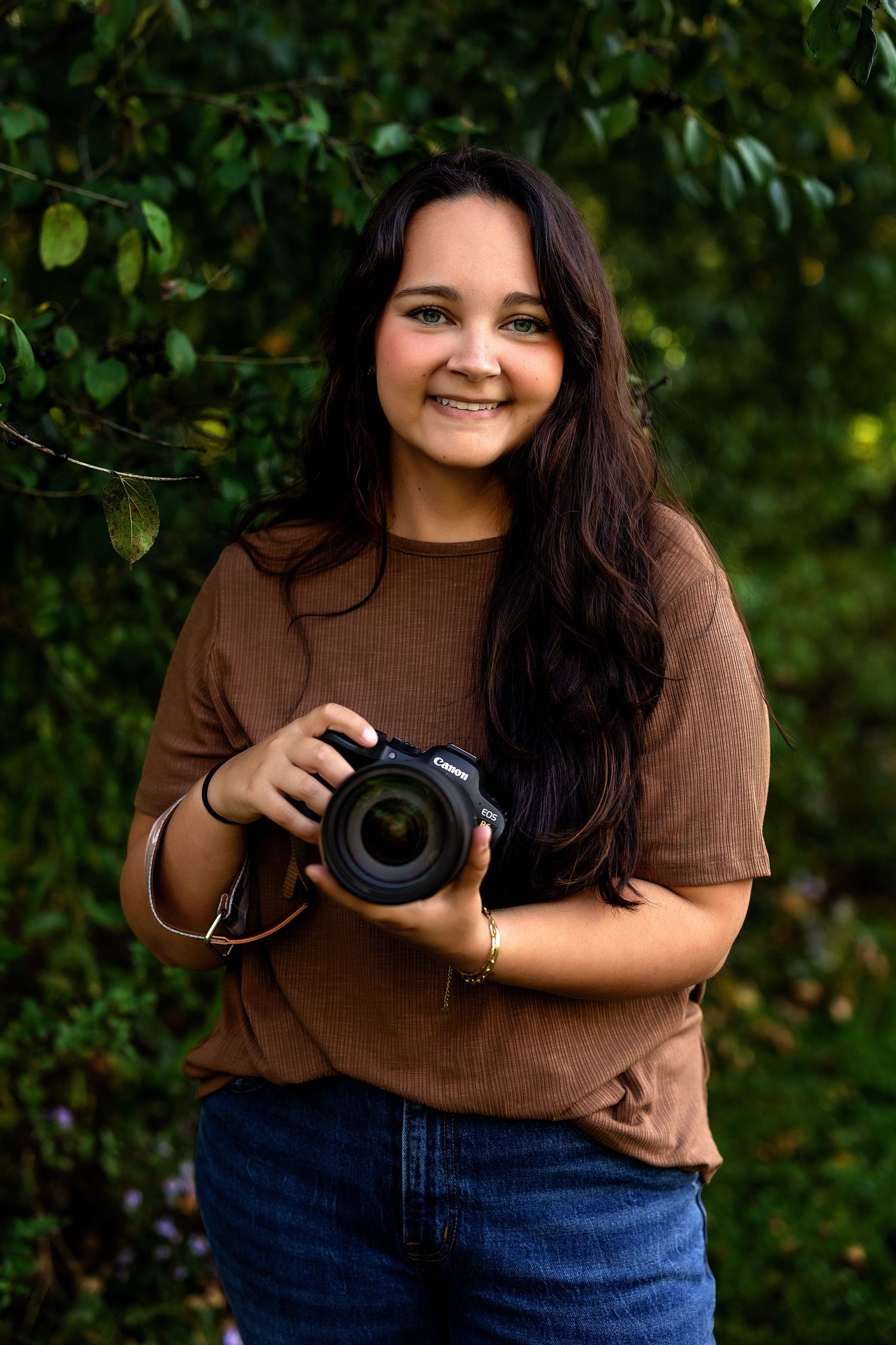 A photographer in a brown shirt and jeans holds a camera while posing outdoors against green foliage.