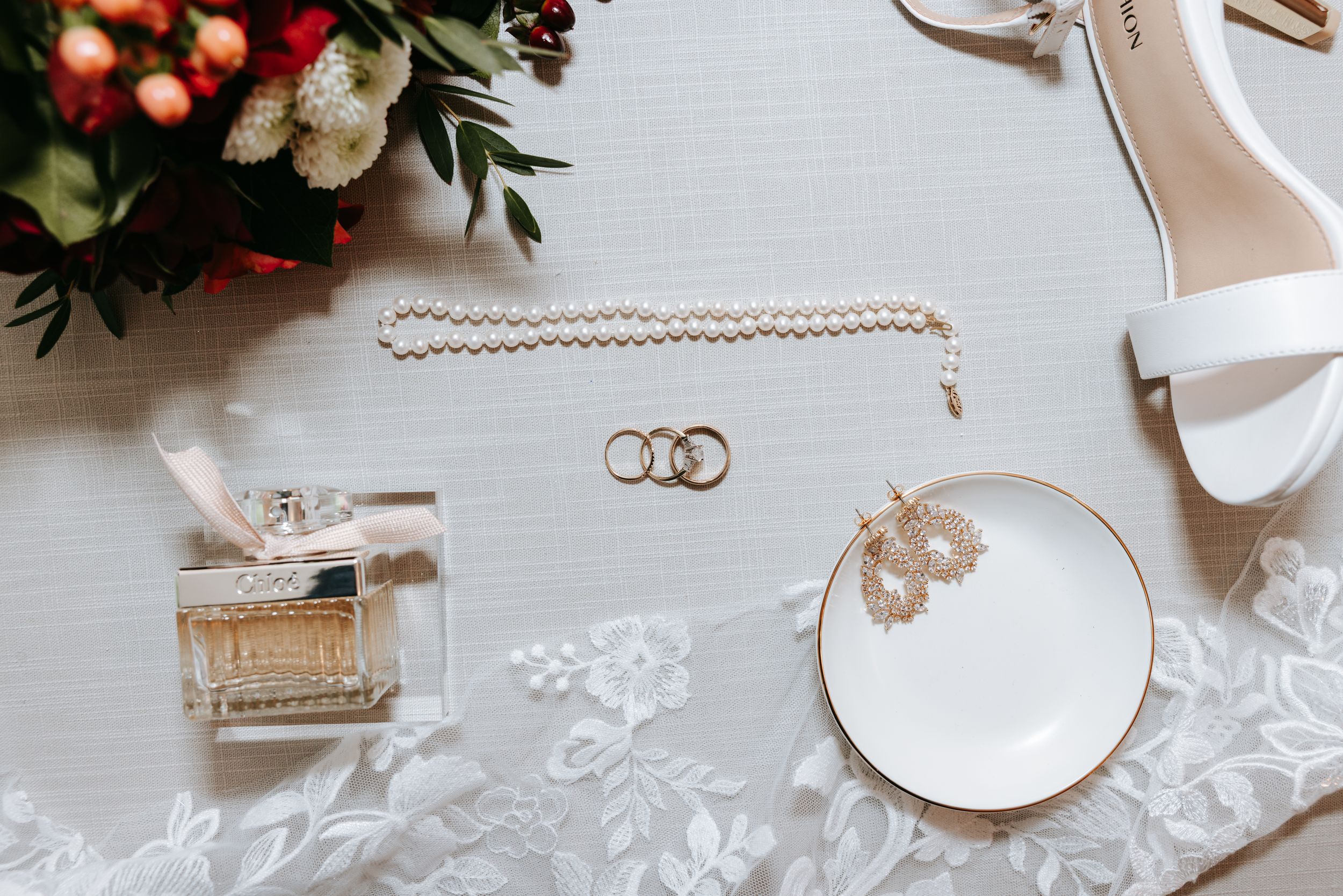 Wedding detail flatlay showing rings, florals and decorative elements on white linen surface.