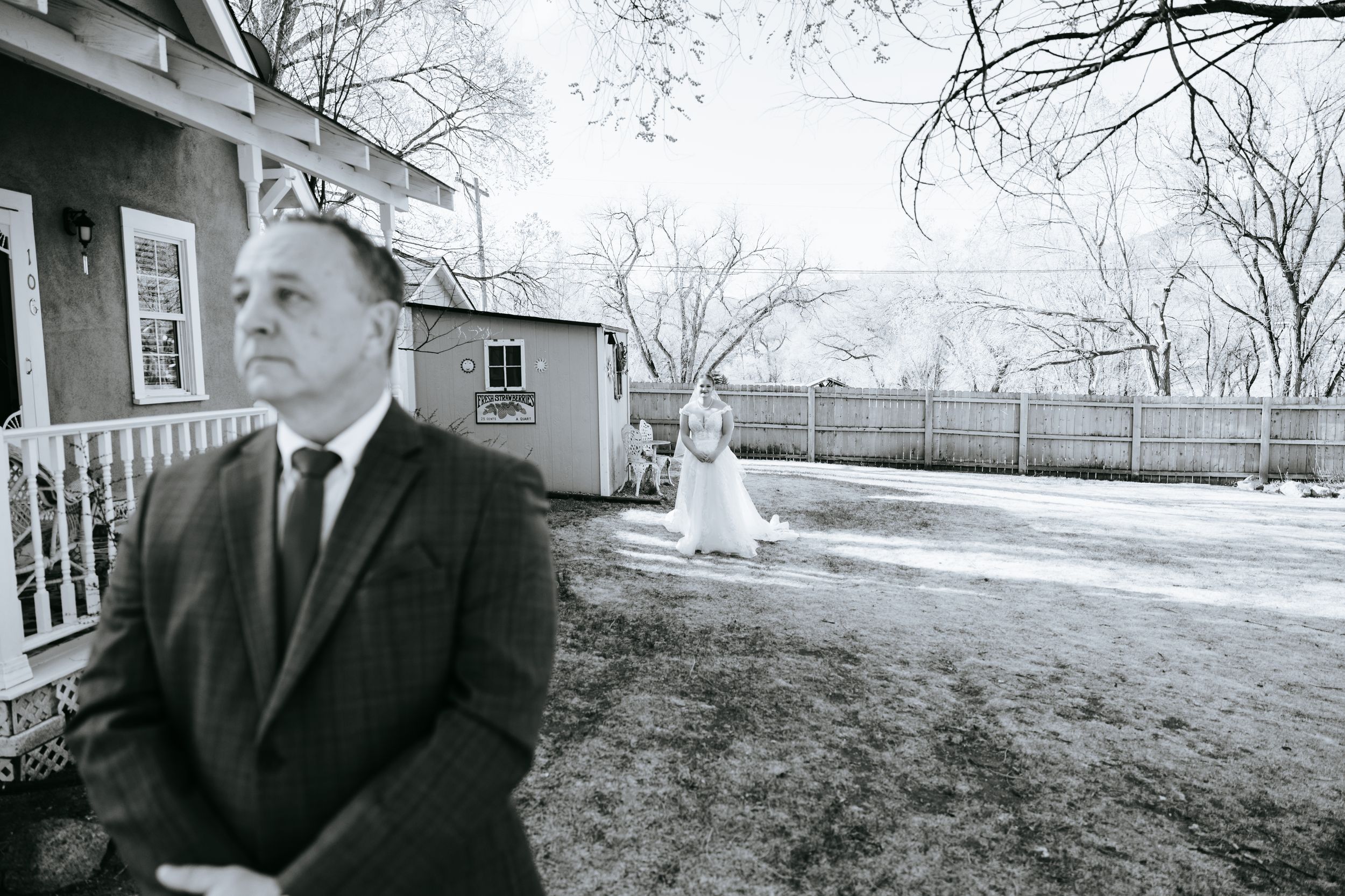 A black and white wedding photo taken in front of a house with bare winter trees.