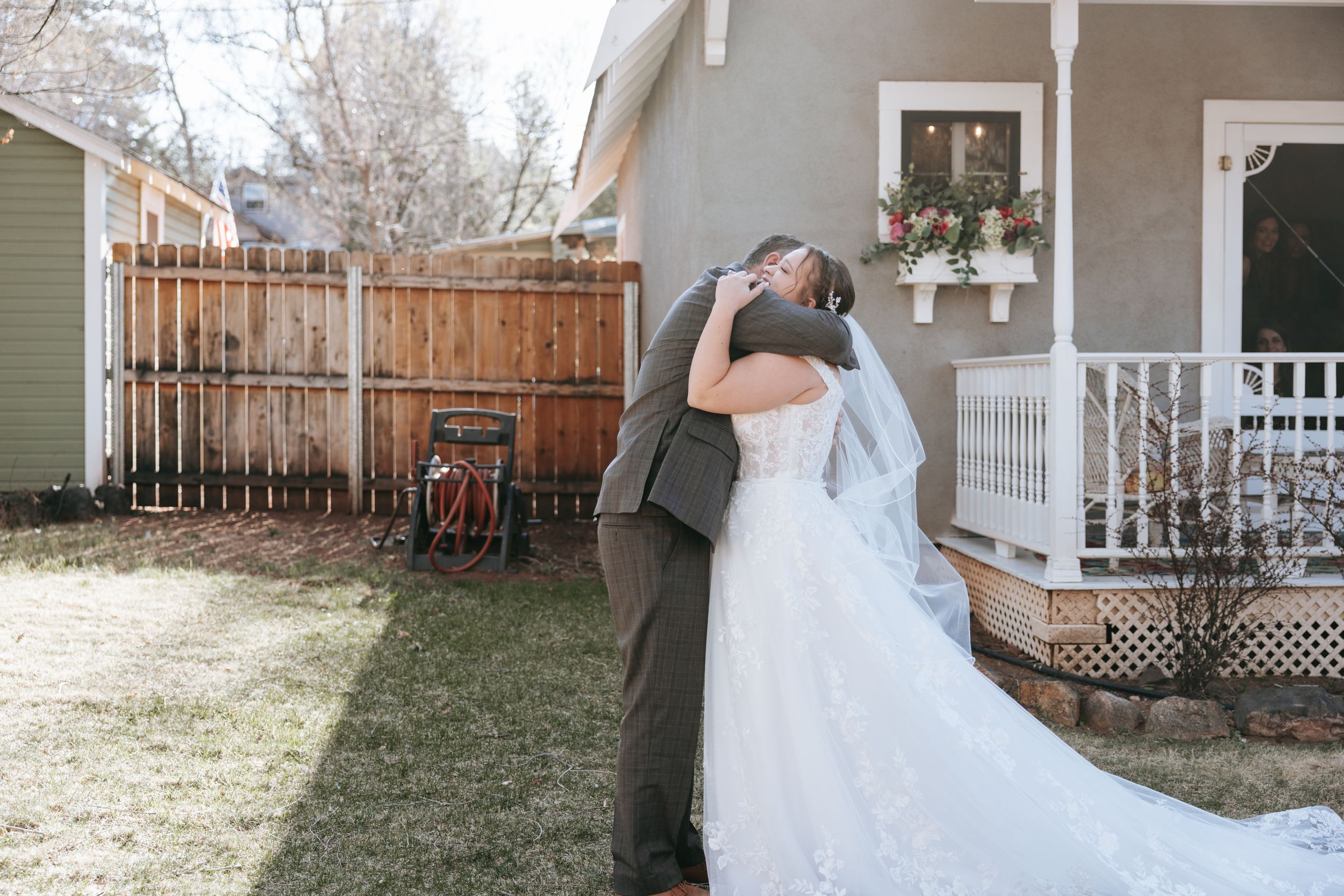 A wedding couple embraces in front of a house with a white porch on a sunny day.