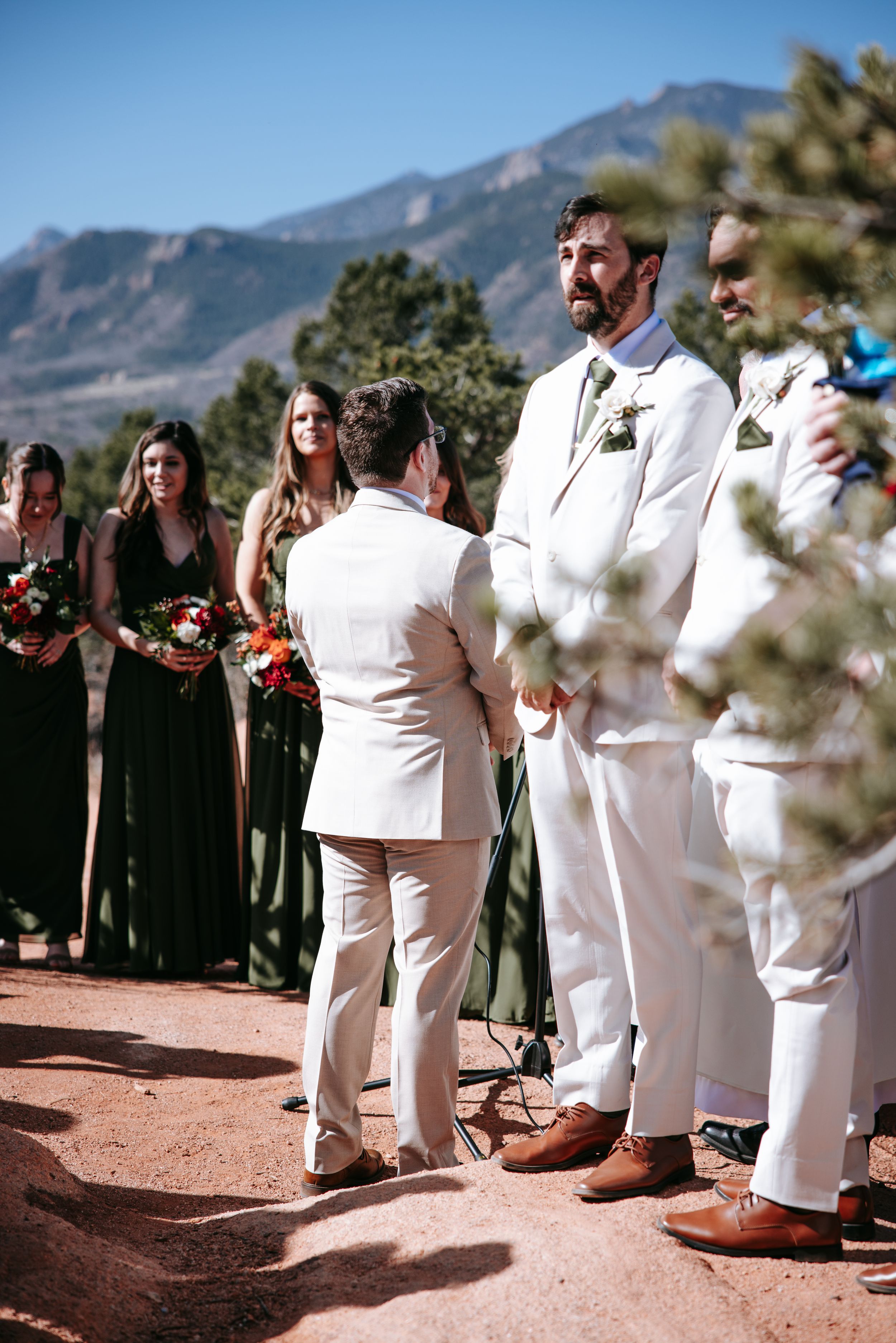 An outdoor wedding ceremony takes place with attendants in white suits and dark dresses against a mountainous backdrop.