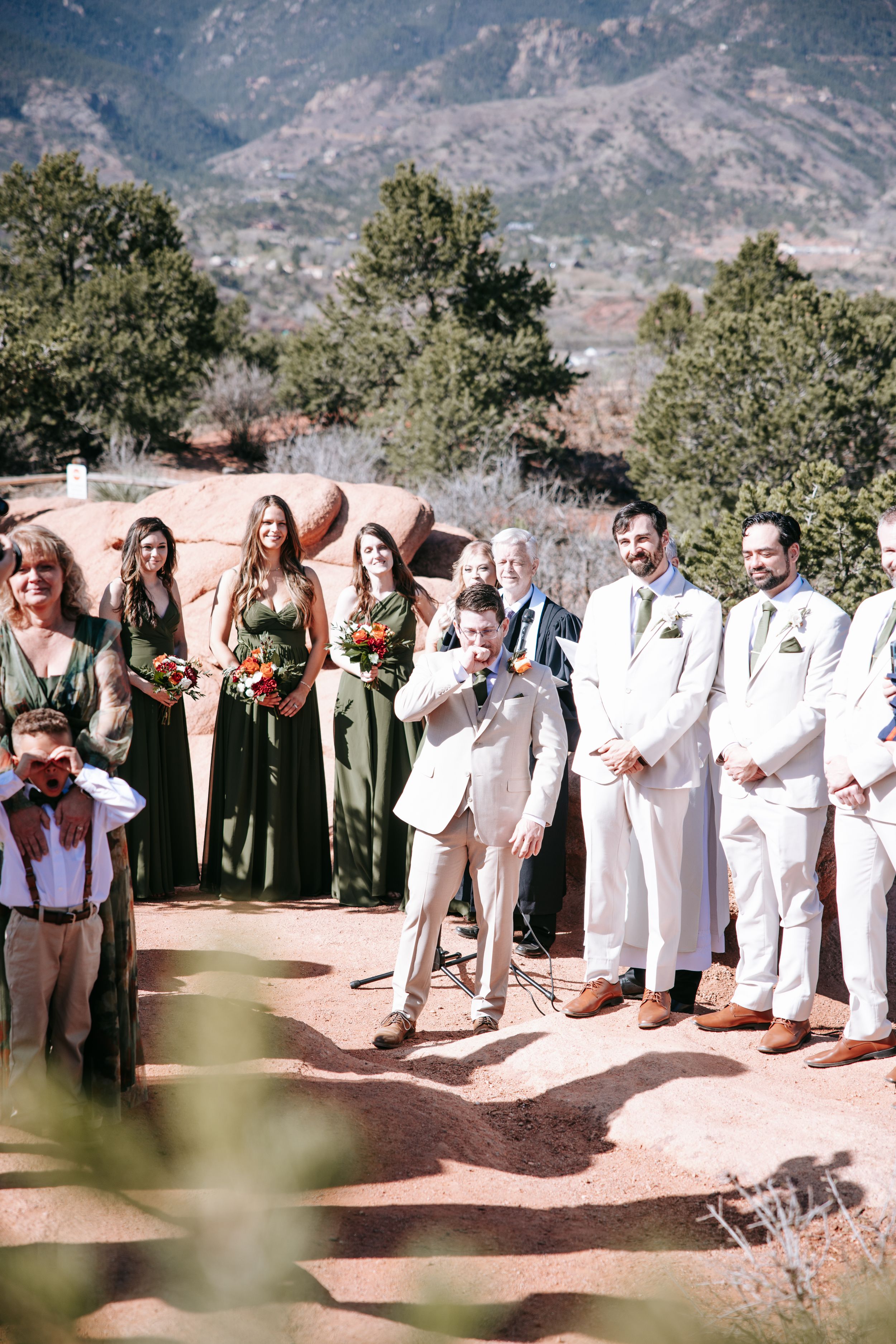 Wedding party in sage green dresses and white suits poses outdoors against mountain backdrop at Garden of the Gods.