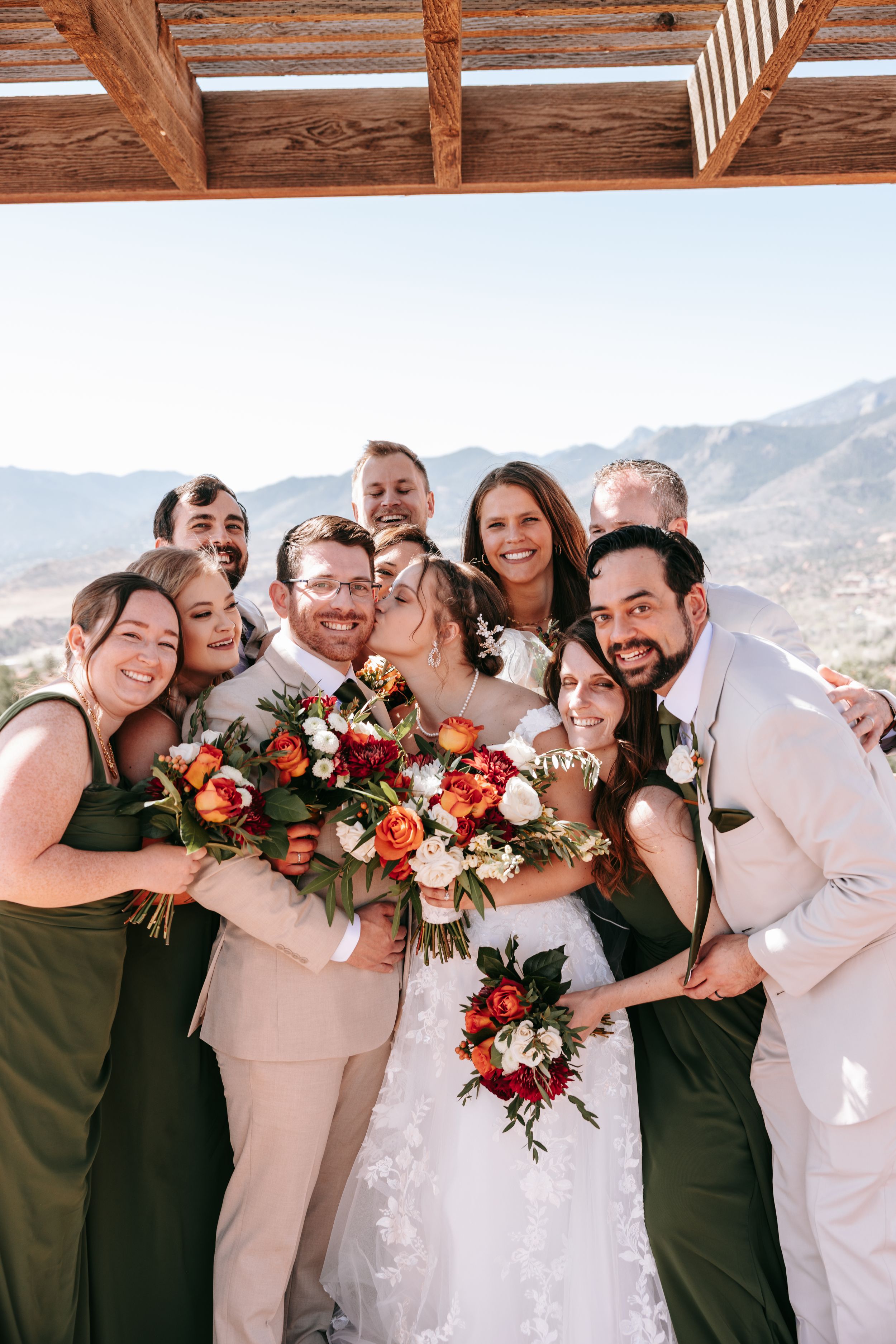 Wedding party holding vibrant red and orange bouquets poses together against mountain backdrop on sunny day.